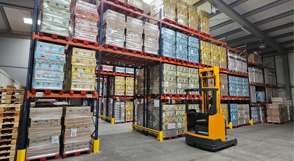 Magazyn z wysokimi regałami magazynowymi wypełnionymi towarami w pudełkach, uporządkowanymi i oznakowanymi. Żółty wózek widłowy jest zaparkowany w alejce, gotowy do użycia. Przestronny, dobrze oświetlony obszar sugeruje niedawny przegląd regałów pod kątem bezpieczeństwa i wydajności.
