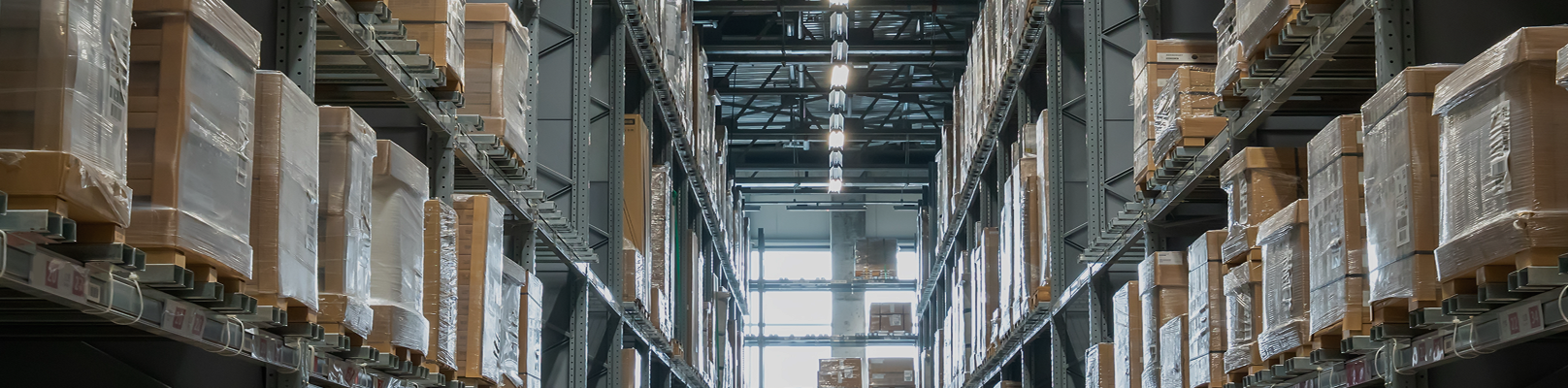 Wnętrze magazynu z wysokimi regałami magazynowymi ułożonymi w stosy z dużymi, termokurczliwymi kartonami po obu stronach i naturalnym światłem wpadającym przez okna na drugim końcu - idealne miejsce na dokładny przegląd regałów.