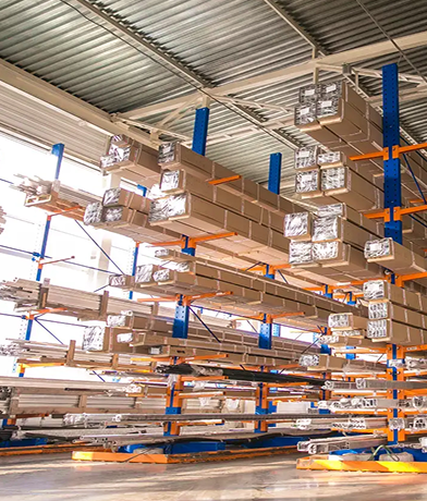 Stosy długich metalowych belek owiniętych plastikiem ułożone są na niebieskich i pomarańczowych regałach magazynowych wewnątrz jasno oświetlonego magazynu z dużymi oknami i wysokim sufitem, gotowego do przeglądu regałów.