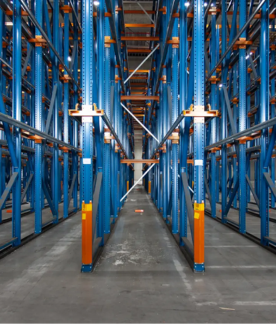 Industrialne wnętrze magazynu z wysokimi niebiesko-pomarańczowymi regałami magazynowymi tworzącymi wąskie korytarze, pustymi półkami i betonową podłogą. Przestrzeń wydaje się zorganizowana, przestronna i dobrze oświetlona - idealna do wydajnego przeglądu regałów.