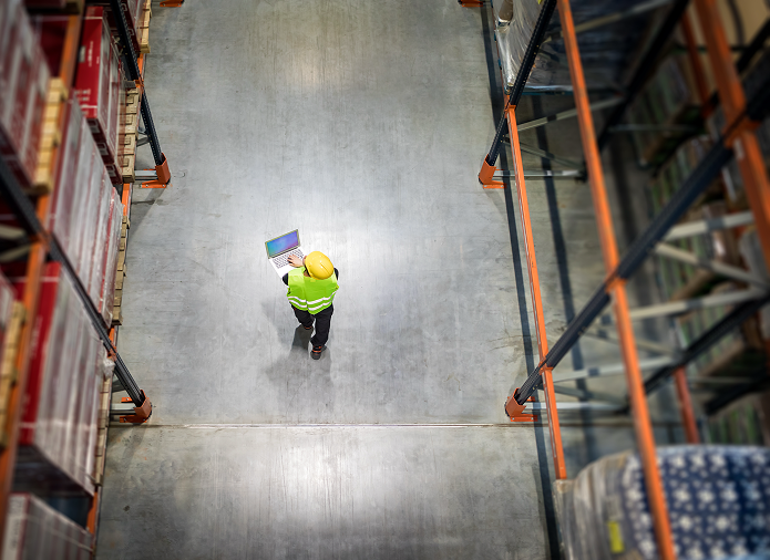 Widok z góry na osobę w żółtym kasku ochronnym i kamizelce odblaskowej idącą korytarzem, trzymającą schowek i wykonującą przegląd regałów wśród regałów magazynowych wypełnionych kartonami.