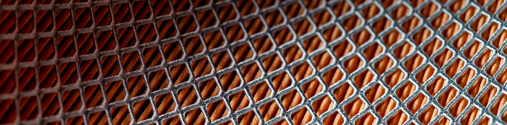 Zbliżenie na srebrną metalową siatkę z otworami w kształcie rombów, odsłaniającymi pomarańczową powierzchnię pod spodem. Wzór przypomina geometryczną, kratkowaną teksturę często spotykaną w regałach magazynowych używanych podczas przeglądu regałów.