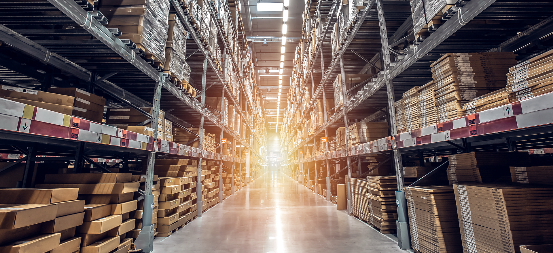 Szeroki korytarz wewnątrz dużego magazynu z wysokimi regałami magazynowymi ułożonymi w stosy kartonowych pudeł i paczek o różnych rozmiarach, oświetlony jasnym światłem słonecznym wpadającym z daleka - idealny do wydajnego przeglądania regałów.