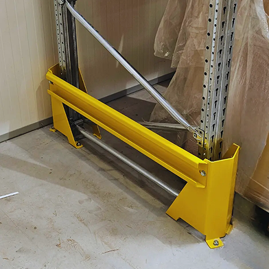 Żółta metalowa osłona jest zainstalowana u podstawy regałów magazynowych, aby chronić je przed uderzeniami. Regał, odpowiedni do regularnego przeglądu regałów, jest umieszczony przy ścianie na betonowej podłodze.
