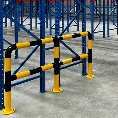 Bariera ochronna w żółto-czarne pasy jest zainstalowana na betonowej posadzce wewnątrz magazynu, obok niebieskich metalowych regałów magazynowych, zapewniając ochronę podczas regularnych przeglądów regałów i codziennych operacji.