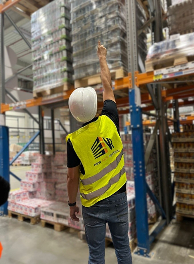 Osoba w żółtej kamizelce ochronnej i białym kasku stoi w magazynie, wskazując w górę na palety ułożone na regałach magazynowych. Pudełka i produkty są ułożone na regałach za nimi podczas przeglądu regałów.