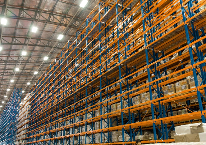 Rzędy wysokich, pomarańczowych i niebieskich regałów magazynowych w dużym magazynie, wypełnione ułożonymi w stos pudełkami i towarami pod jasnym oświetleniem górnym, oferują zorganizowane przechowywanie i łatwy przegląd regałów do zarządzania zapasami.